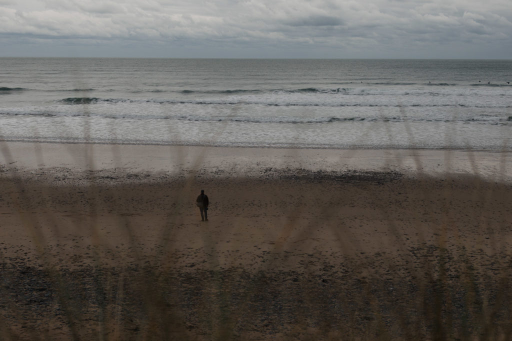 Image of a man in shadow alone on a beach with a wide expanse of waves. He is visible through a fortground of grasses. Image used to represent books catagorised as being that re quiet and reflective