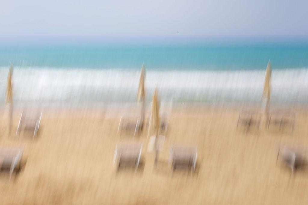 An intentional camera movement photo of sunbeds and parasols against a vivide blue sea. The blur is vertical. Image used to represent books catagorised as being about joyful escapism