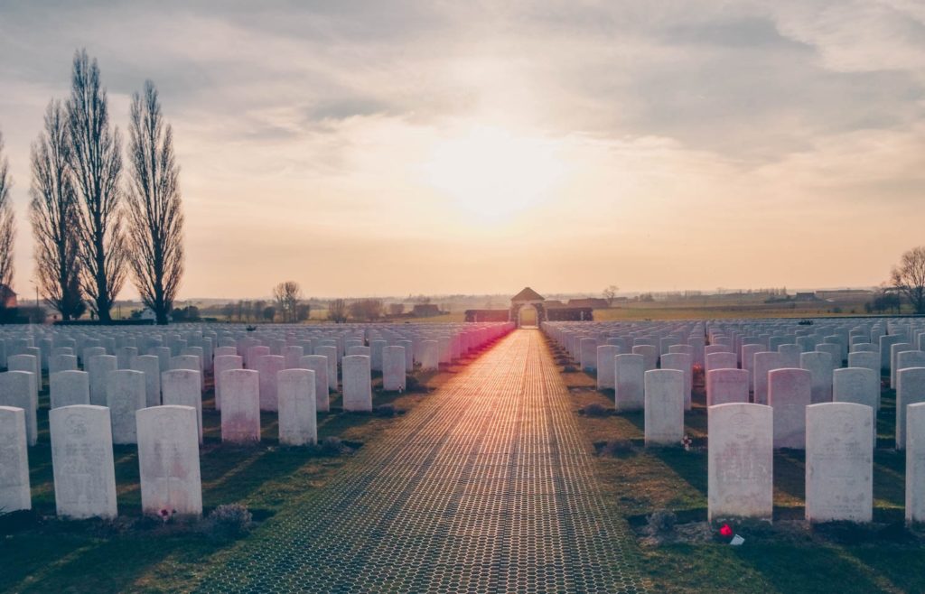 A photo of late night sun shining on the gate and path at Tyne Cot war memorial. On either side white portland stone graves. 