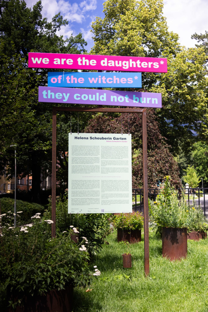 Photo of a pink and purple sign saying 'we are the daughters of the wtches they could not burn'. It is set in a garden in Insbrook with memorial text about Helena below.